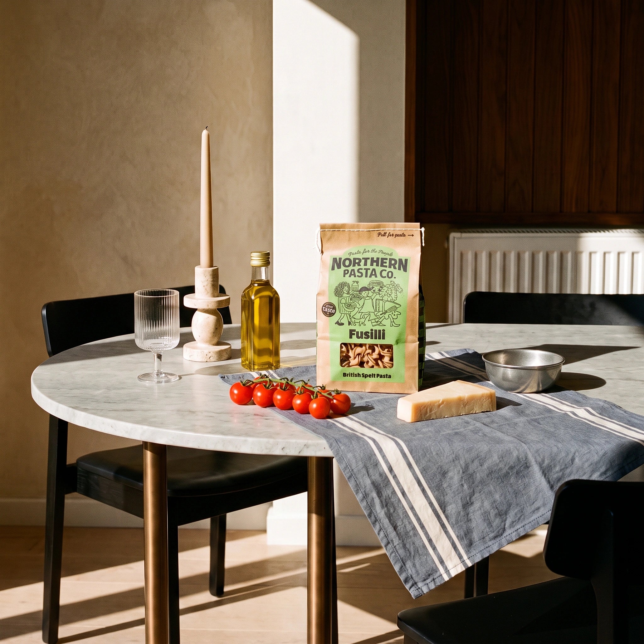 Northern Pasta Co. British Spelt Fusilli bag on a sunlit marble dining table with olive oil, tomatoes and parmesan