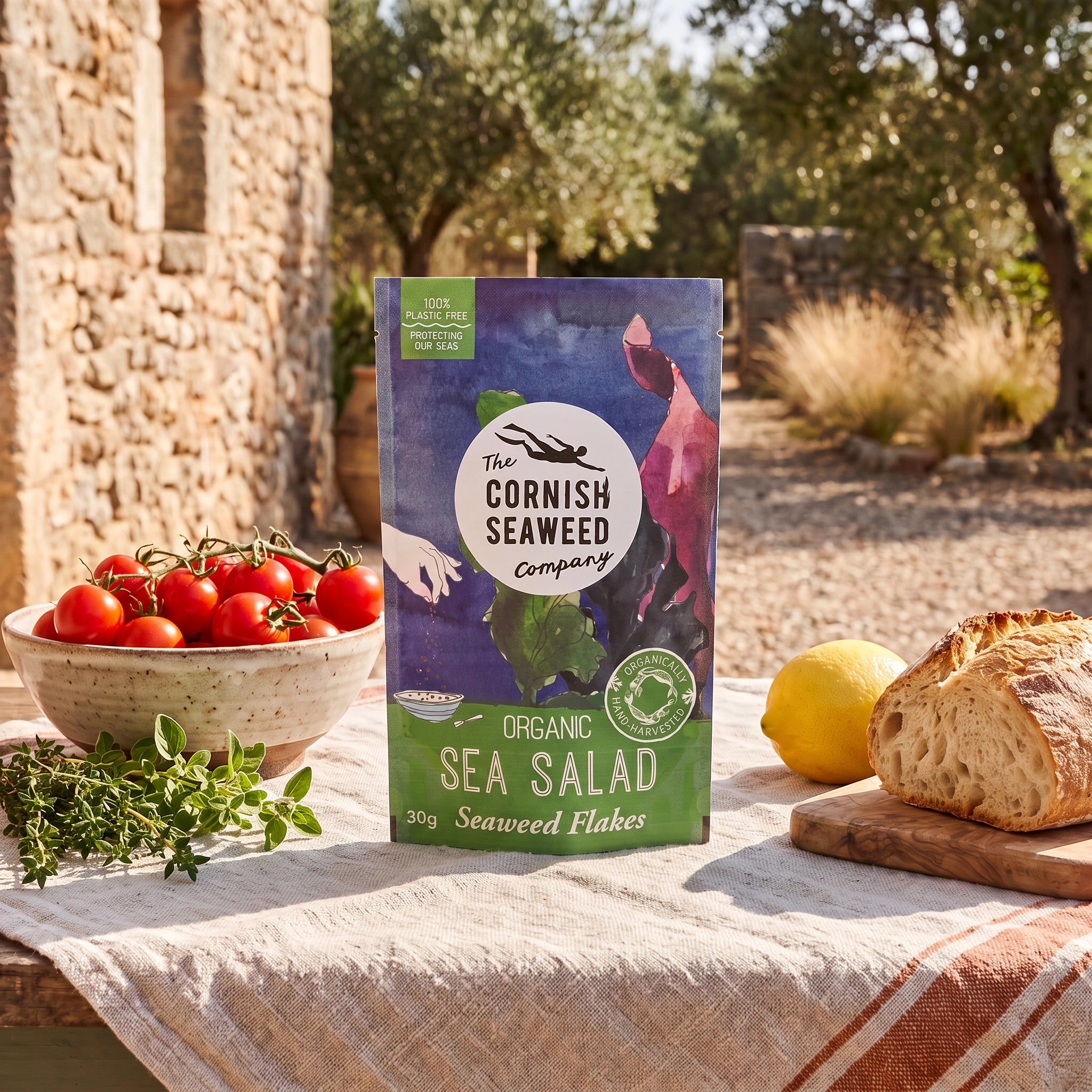 The Cornish Seaweed Company Organic Sea Salad Flakes pouch on a sunlit patio table in an olive grove with tomatoes and herbs