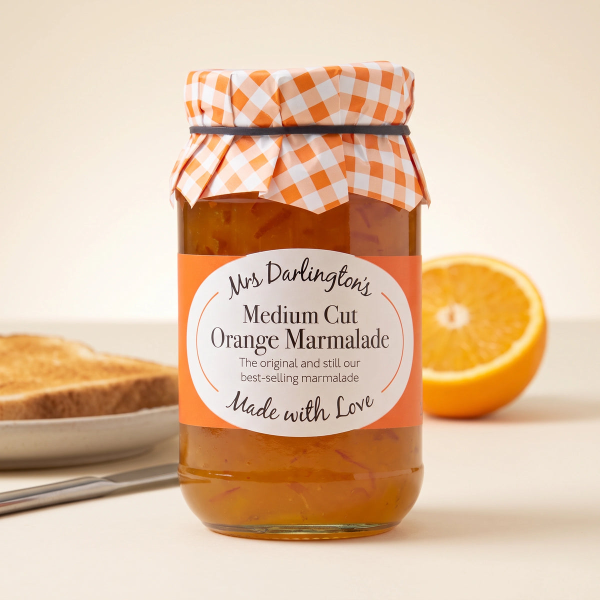 Mrs Darlington's Medium Cut Orange Marmalade jar on a pale table with buttered toast, a knife and a halved orange