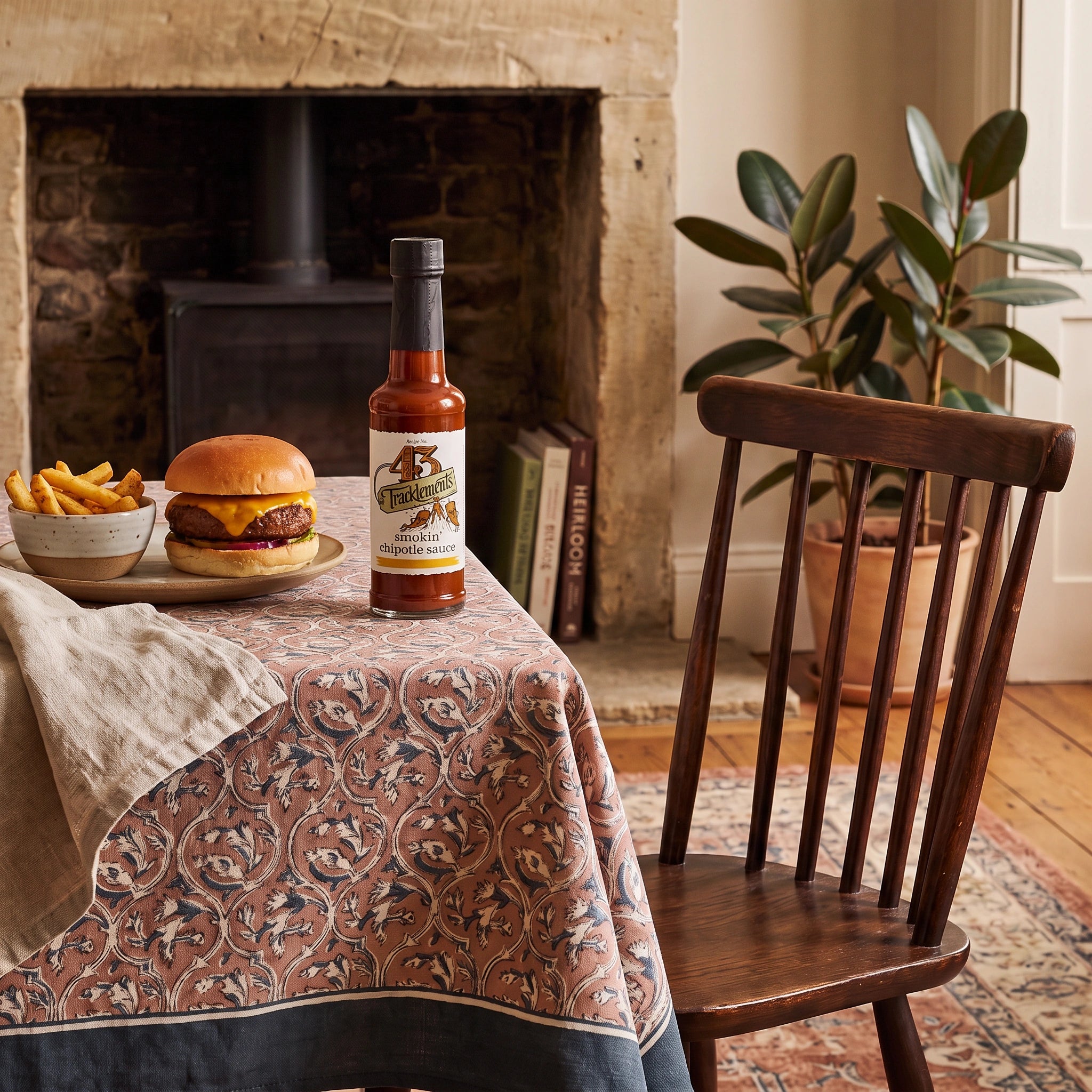 Tracklements Smokin' Chipotle Sauce bottle on a table by a fireplace with a cheeseburger and fries