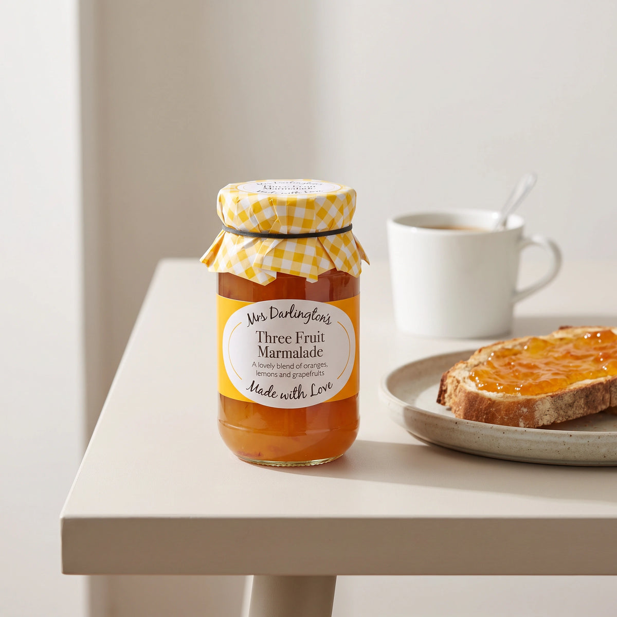 Mrs Darlington's Three Fruit Marmalade jar on a pale table with marmalade on toast and a cup of tea